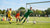 Een jonge keeper traint in een doel, terwijl een coach aanwijzingen geeft met een voetbal in de achtergrond.