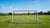 Een verplaatsbaar voetbaldoel staat scheef in de wind, met ballen die rond het doel rollen. Achtergrond met grasveld.